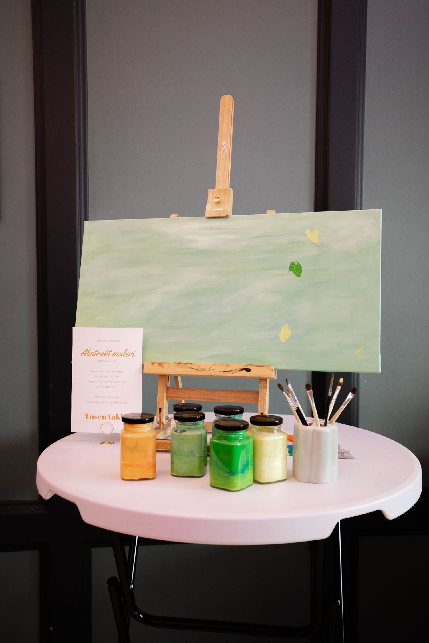 Art supplies and painting materials arranged on a pink table with a green canvas backdrop.