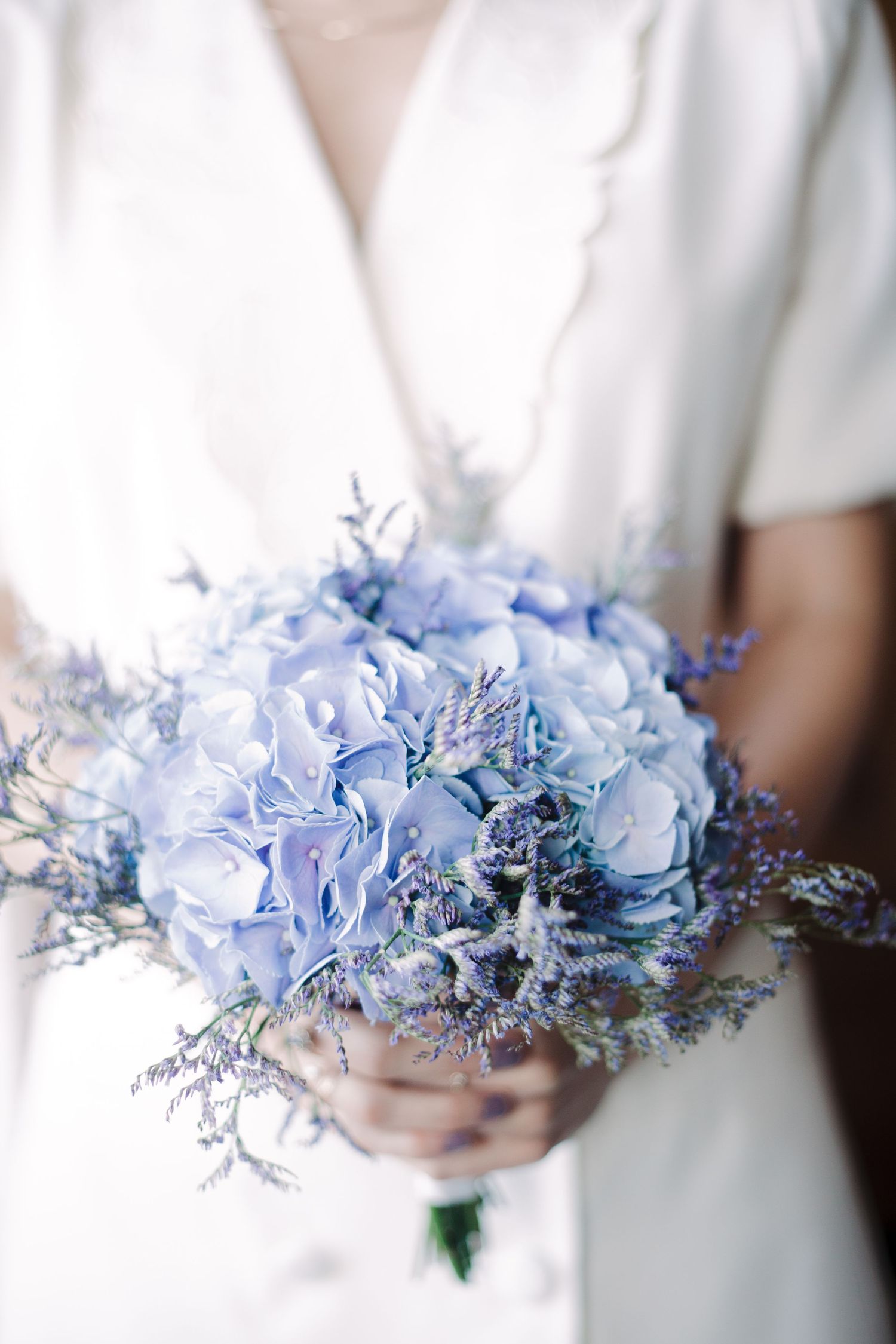 Delicate bouquet of light blue hydrangeas and dried flowers held against a white dress.
