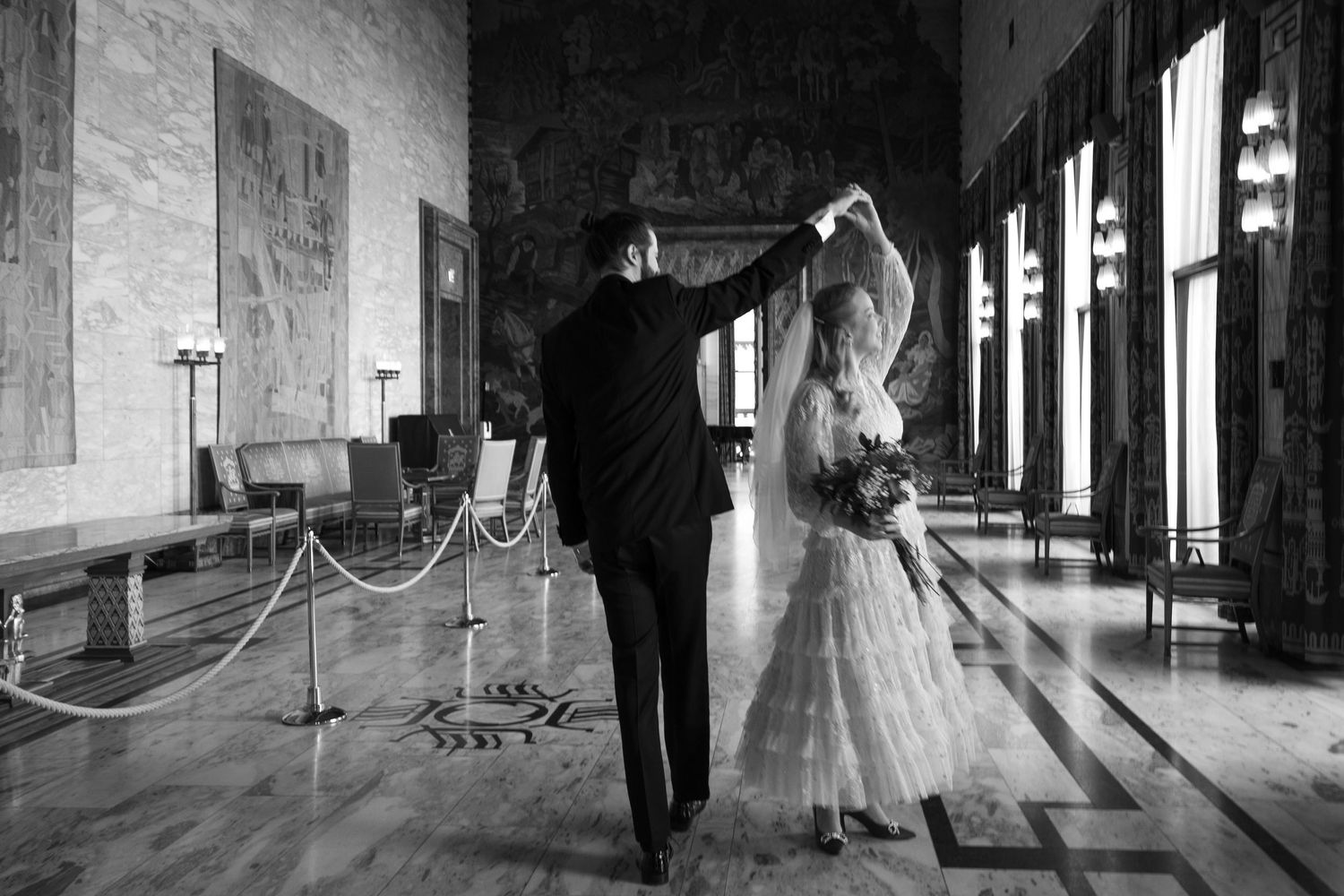 Black and white photo of bride and groom dancing elegantly in a grand hall with tall columns.