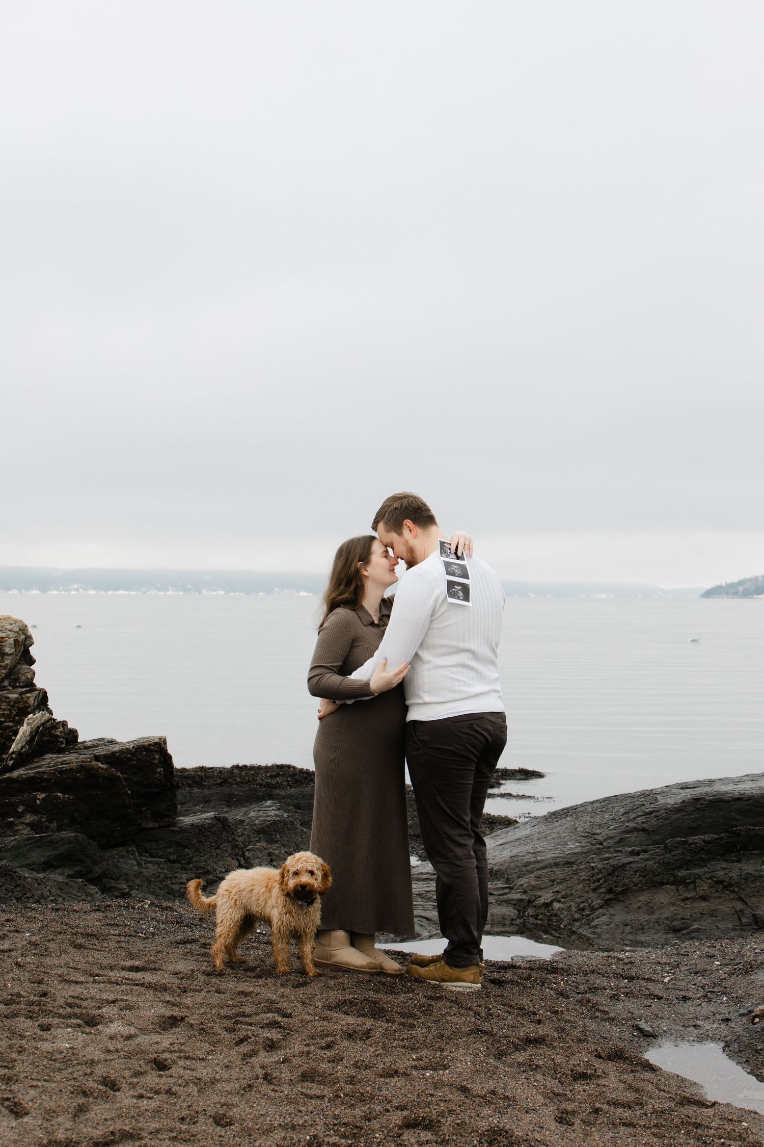 A couple shares a tender moment on rocky coastal cliffs with their golden retriever dog beside them.