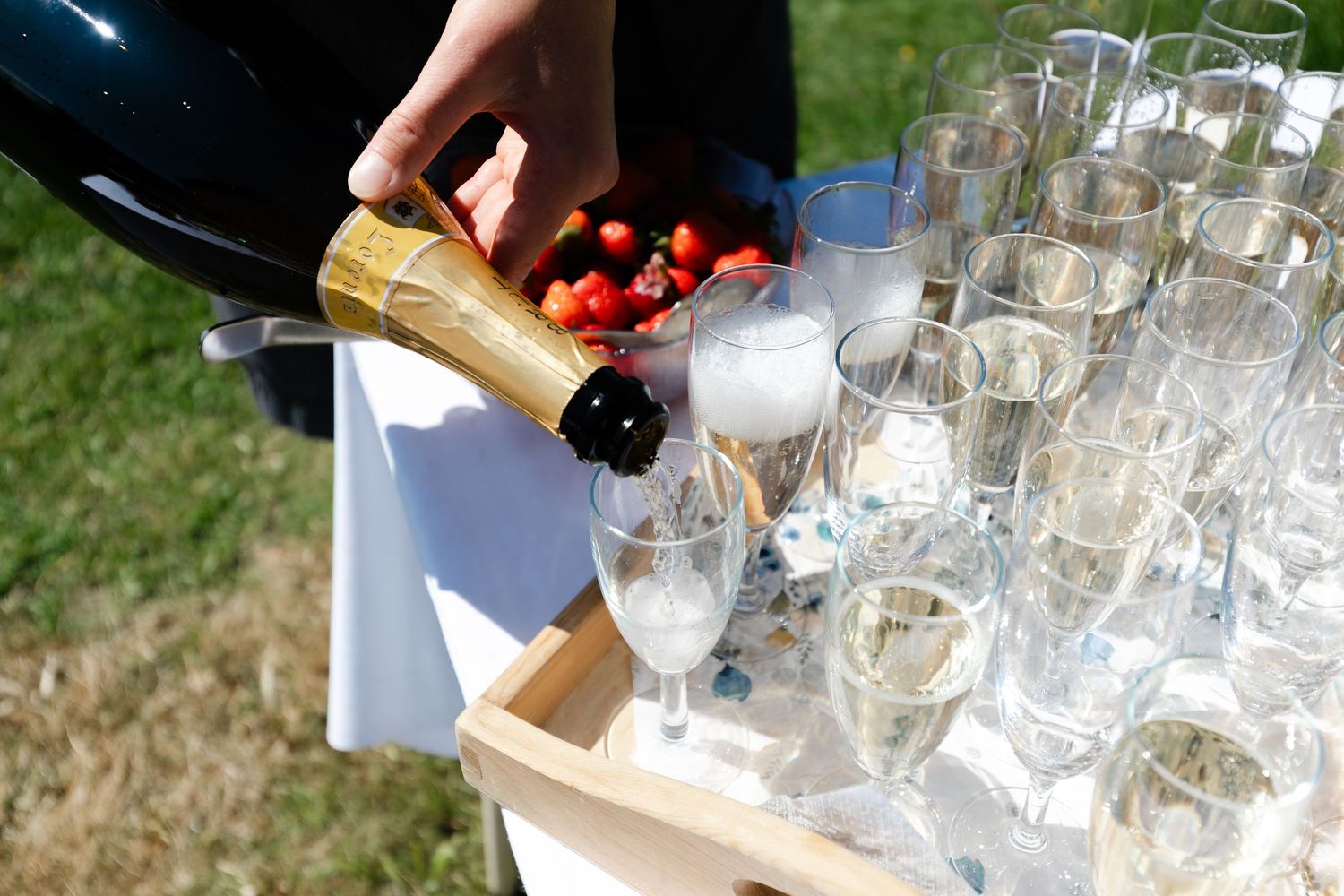 Hands pouring drinks into multiple clear glasses arranged on a wooden surface during an outdoor event.