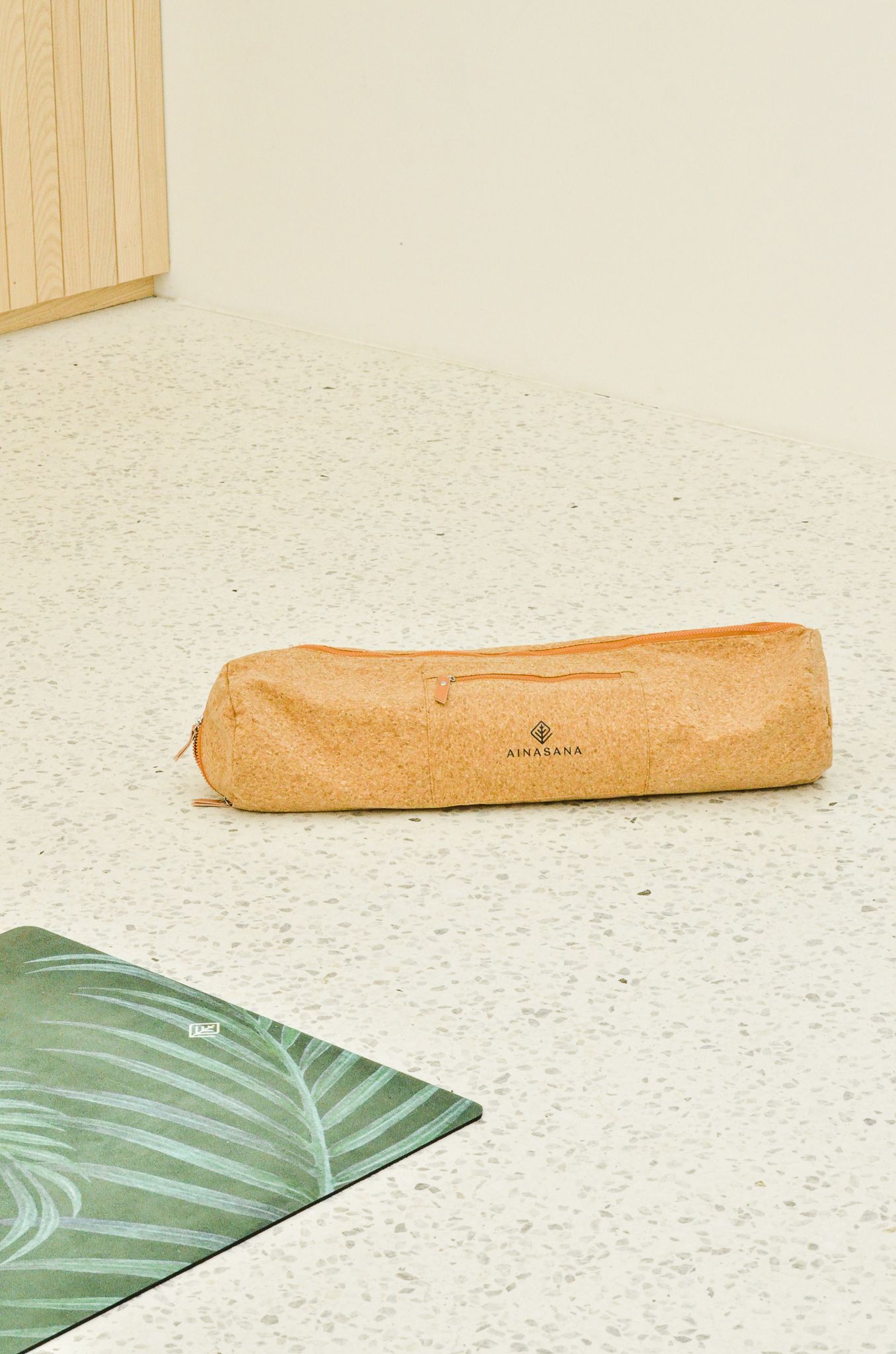 A cork yoga mat bag rests on a white speckled floor next to a green leaf-patterned yoga mat.