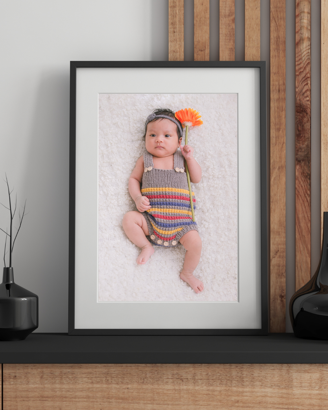 Three framed baby portraits displayed on a wooden wall shelf with decorative black vases.