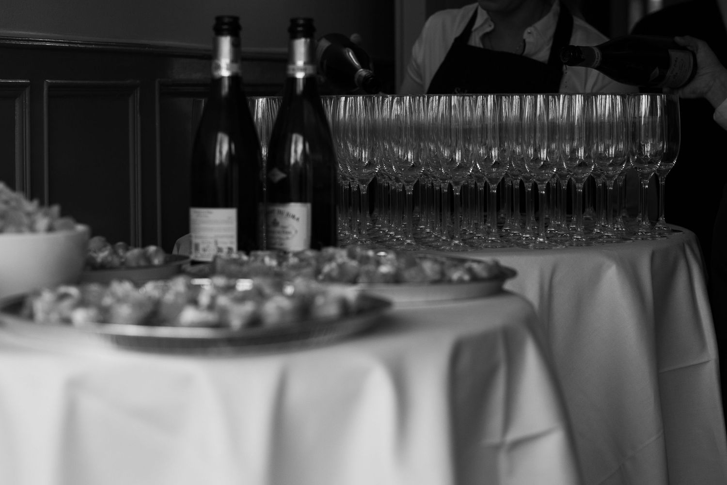 Black and white photo of wine bottles, crystal glasses, and appetizers on a white tablecloth at an elegant event.