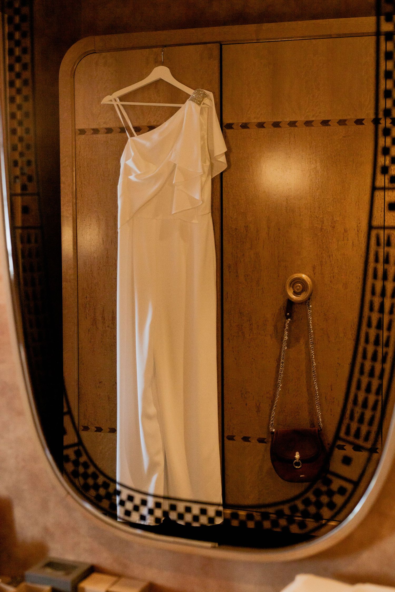 Elegant white one-shoulder wedding dress hanging in a wooden framed mirror.
