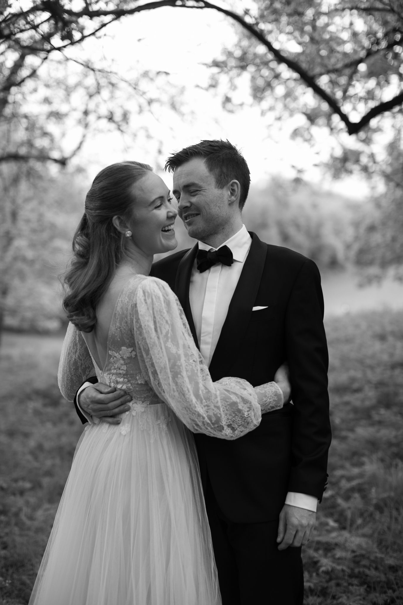 Black and white photo of a bride and groom embracing and smiling outdoors under trees.