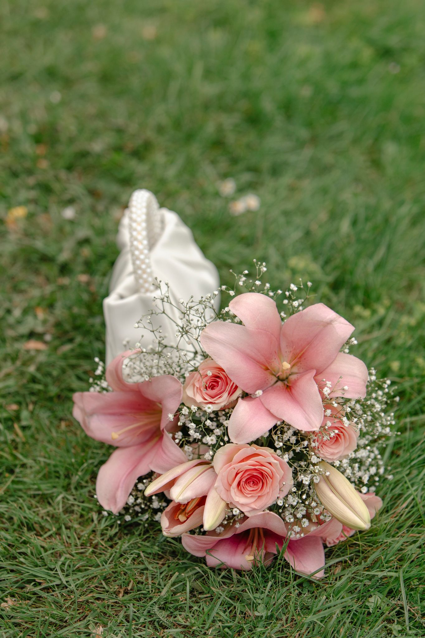 Pink roses and lilies bridal bouquet with baby's breath beside a white satin pearl-handled purse on green grass.