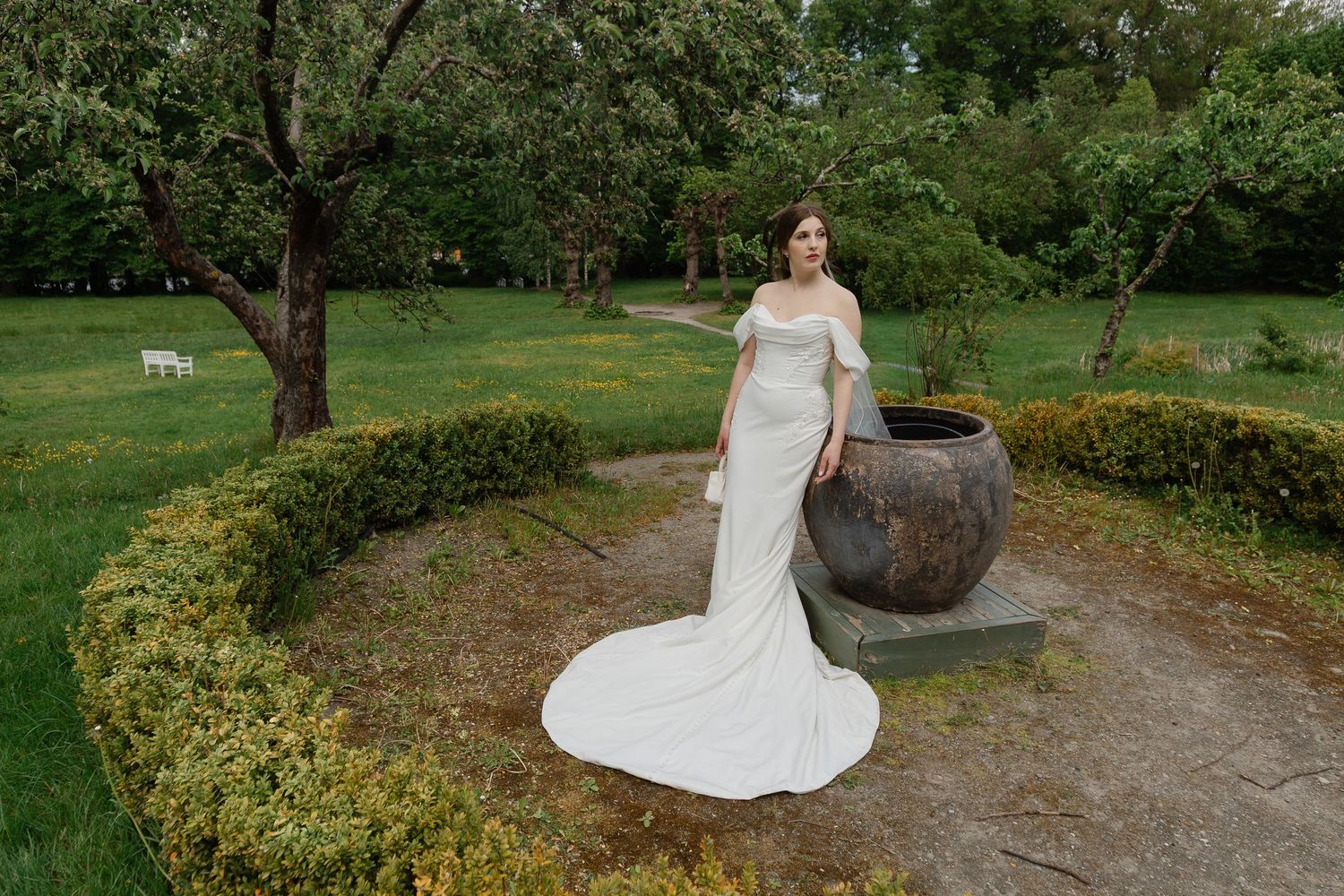 Bride in off-shoulder white gown poses beside large clay pot in lush garden setting.
