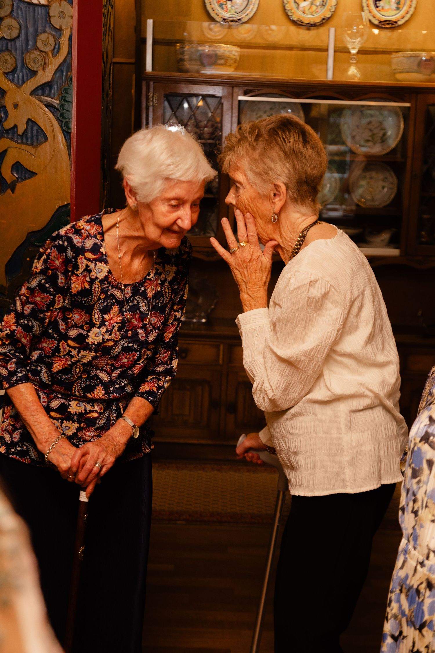 Two elderly women sharing a private conversation and laughing together in front of a wooden cabinet with decorative plates.