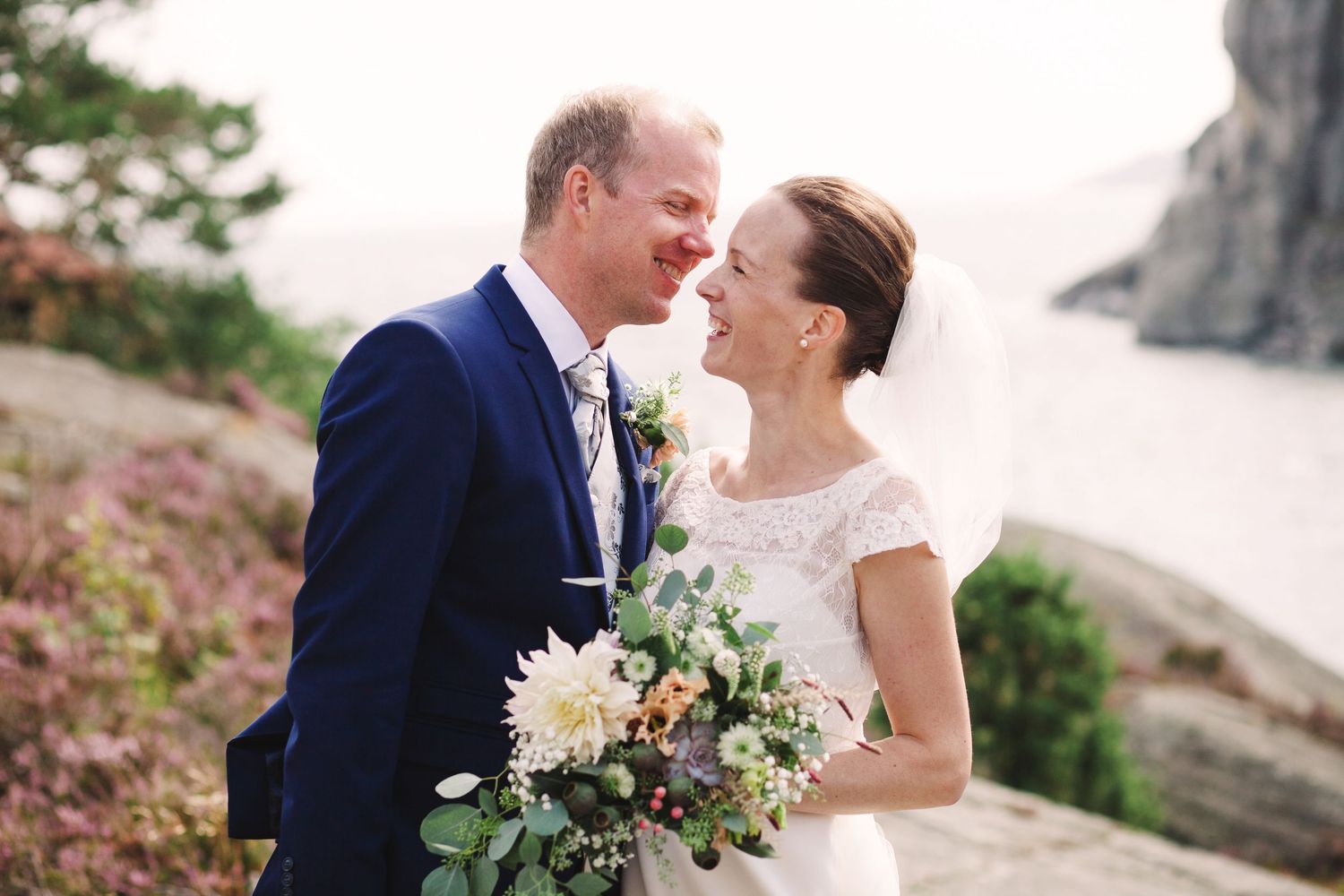 Romantic wedding couple embracing near rocky cliffs with bride holding a pastel floral bouquet.
