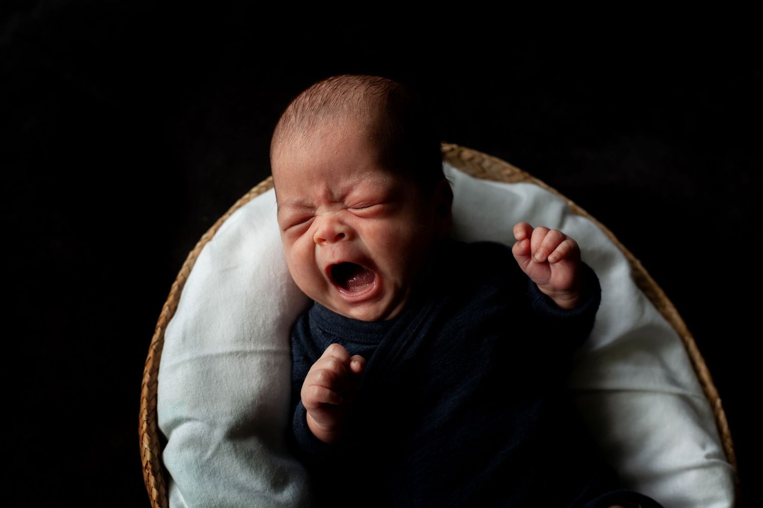 Crying newborn in dark clothing against black background.
