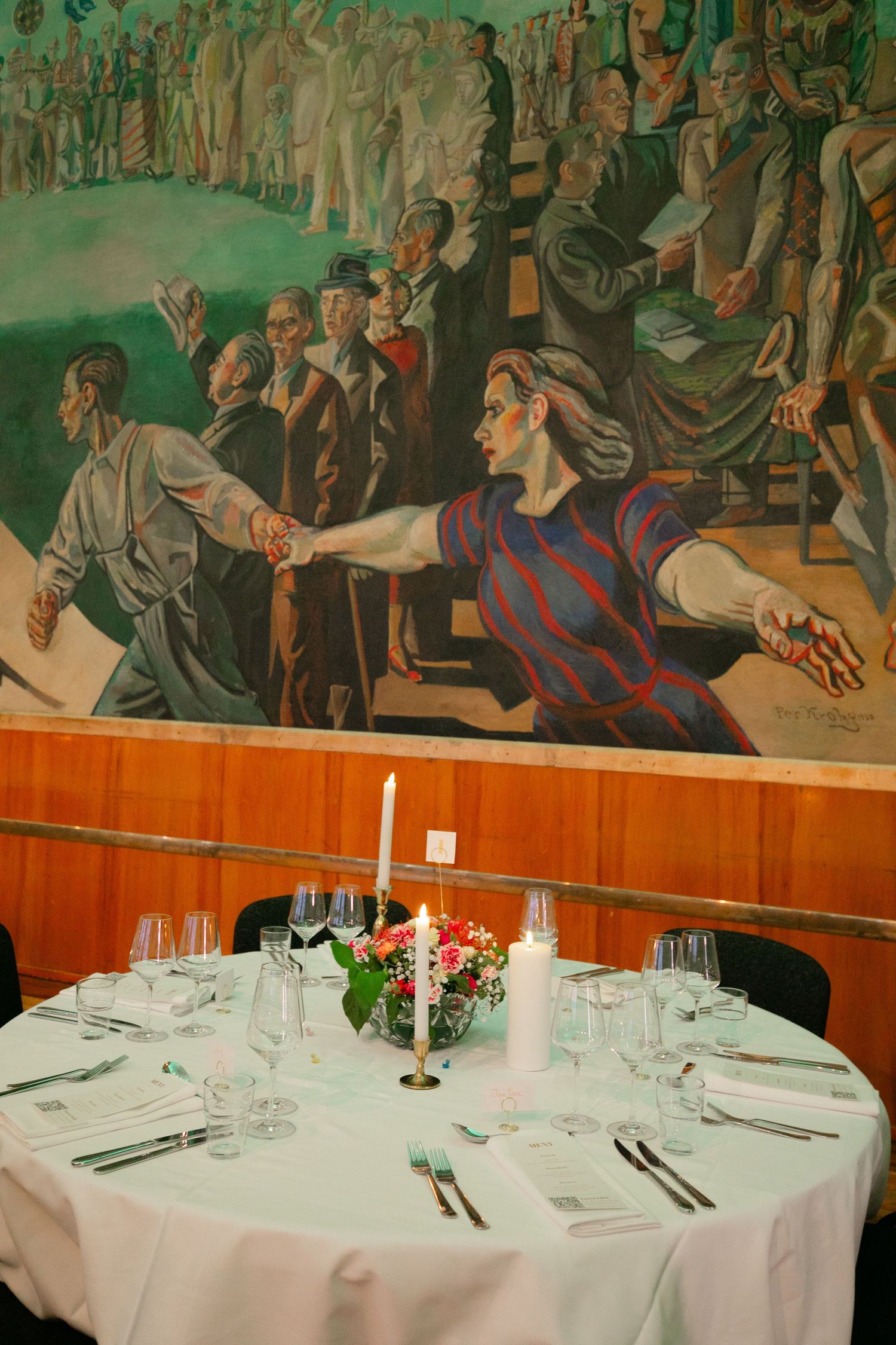 Formal dining table set with white linens and candles against a wall with classical murals.
