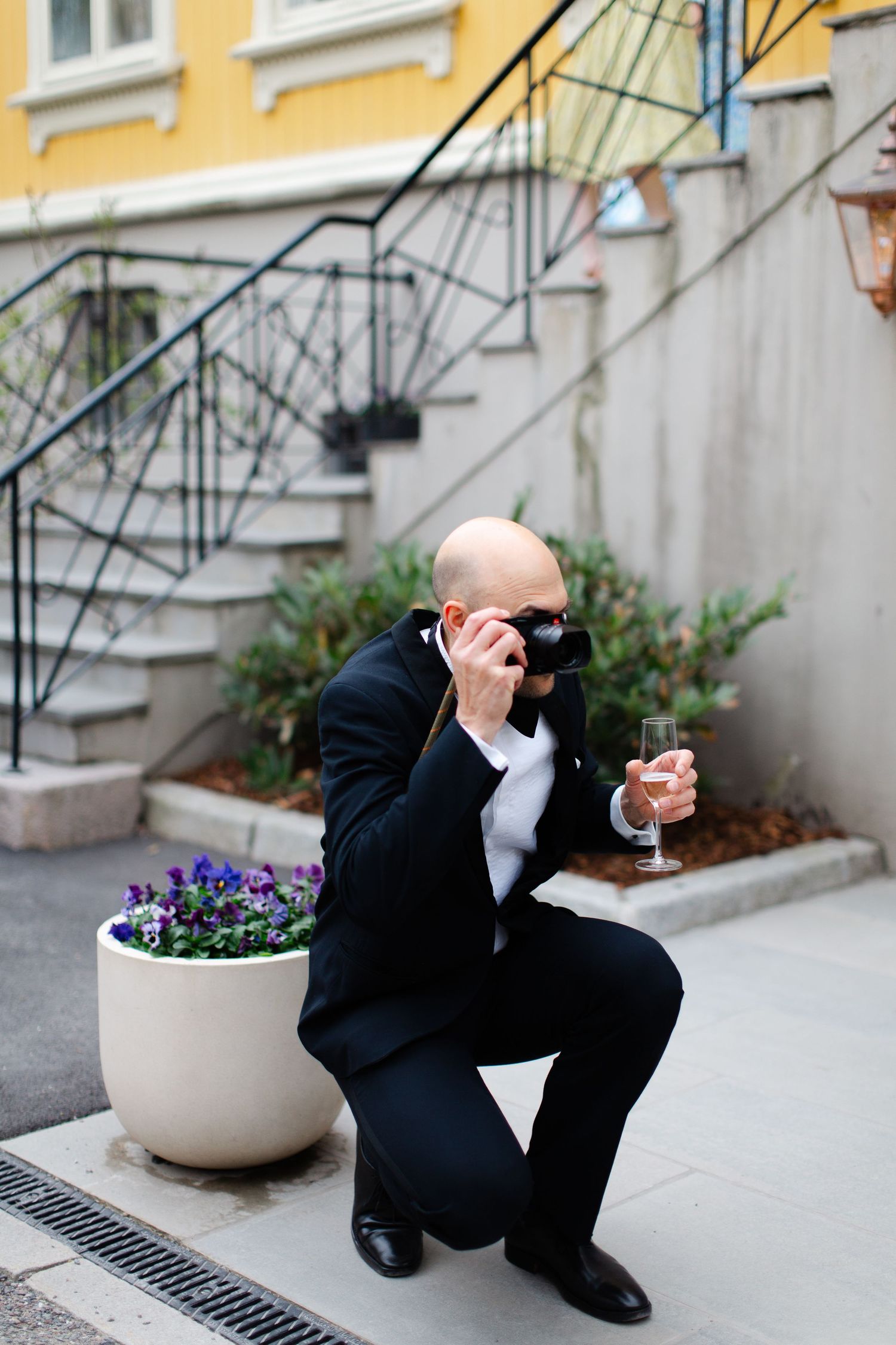 Bald man in black tuxedo crouching outdoors, holding champagne glass while taking photo with camera near flower pot.