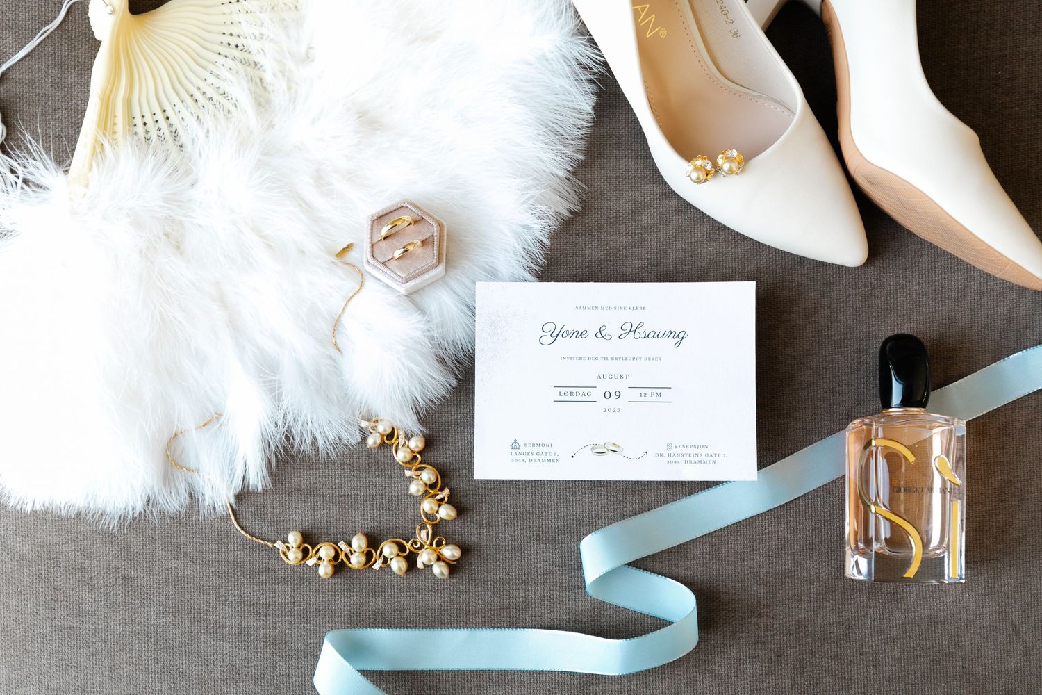 Wedding accessories including white shoes, jewelry, and invitation laid out on grey fabric.