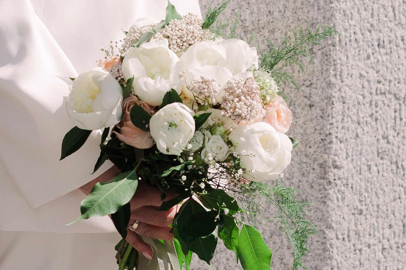 Close-up of a lush white peony and greenery bridal bouquet against a textured wall.