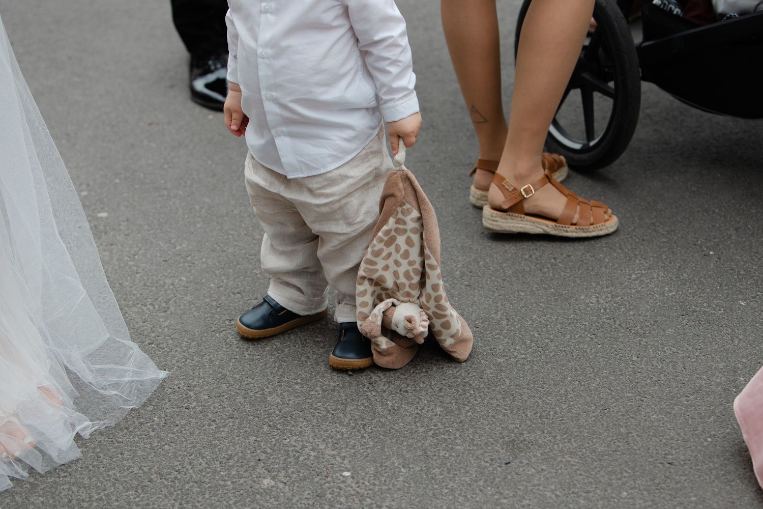 Young child holding a stuffed giraffe toy while standing on pavement at a wedding event.