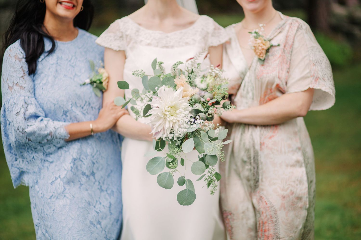 Three women in pastel lace dresses stand together at a wedding with a white and green eucalyptus bouquet.