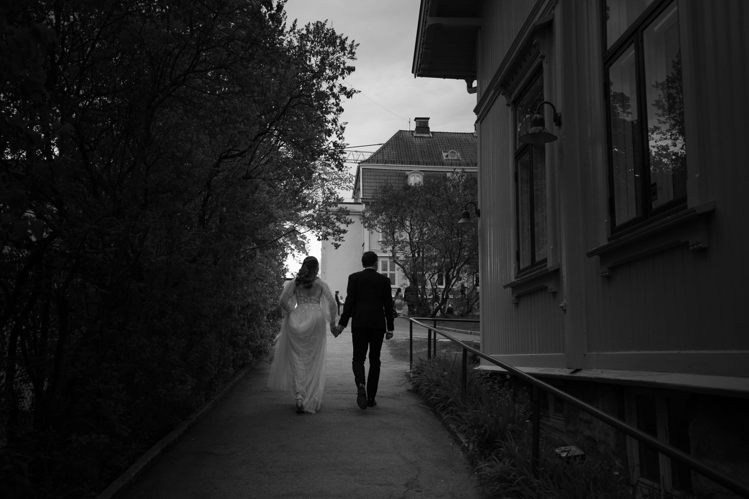 Black and white photo of a bride and groom walking hand in hand down a path beside a building.