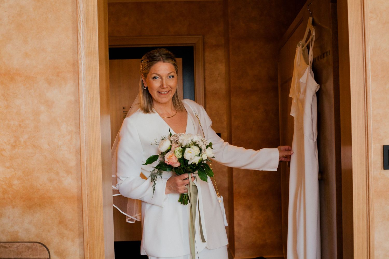 A bride in a white dress stands in a warmly lit hallway holding flowers.