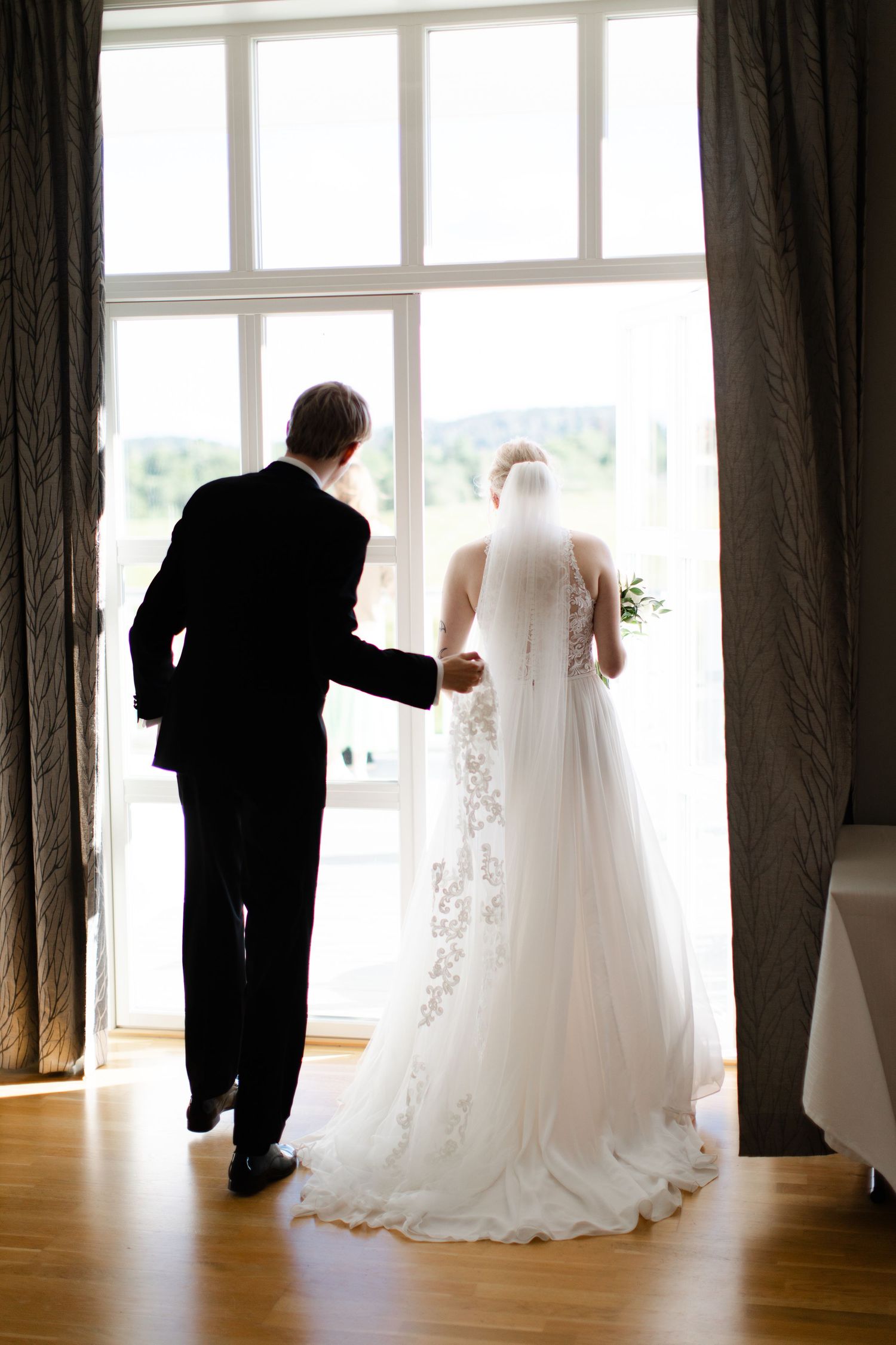 Bride and groom stand hand in hand before large window, viewed from behind, wedding dress with floral lace detail.