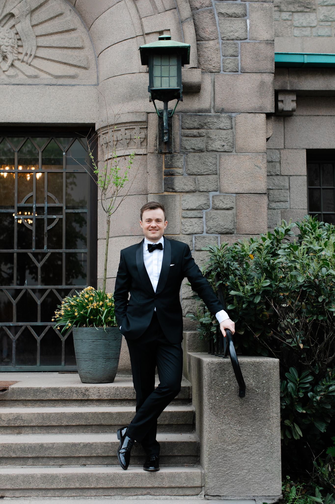 Groom in black tuxedo posing on stone steps outside a historic stone building.