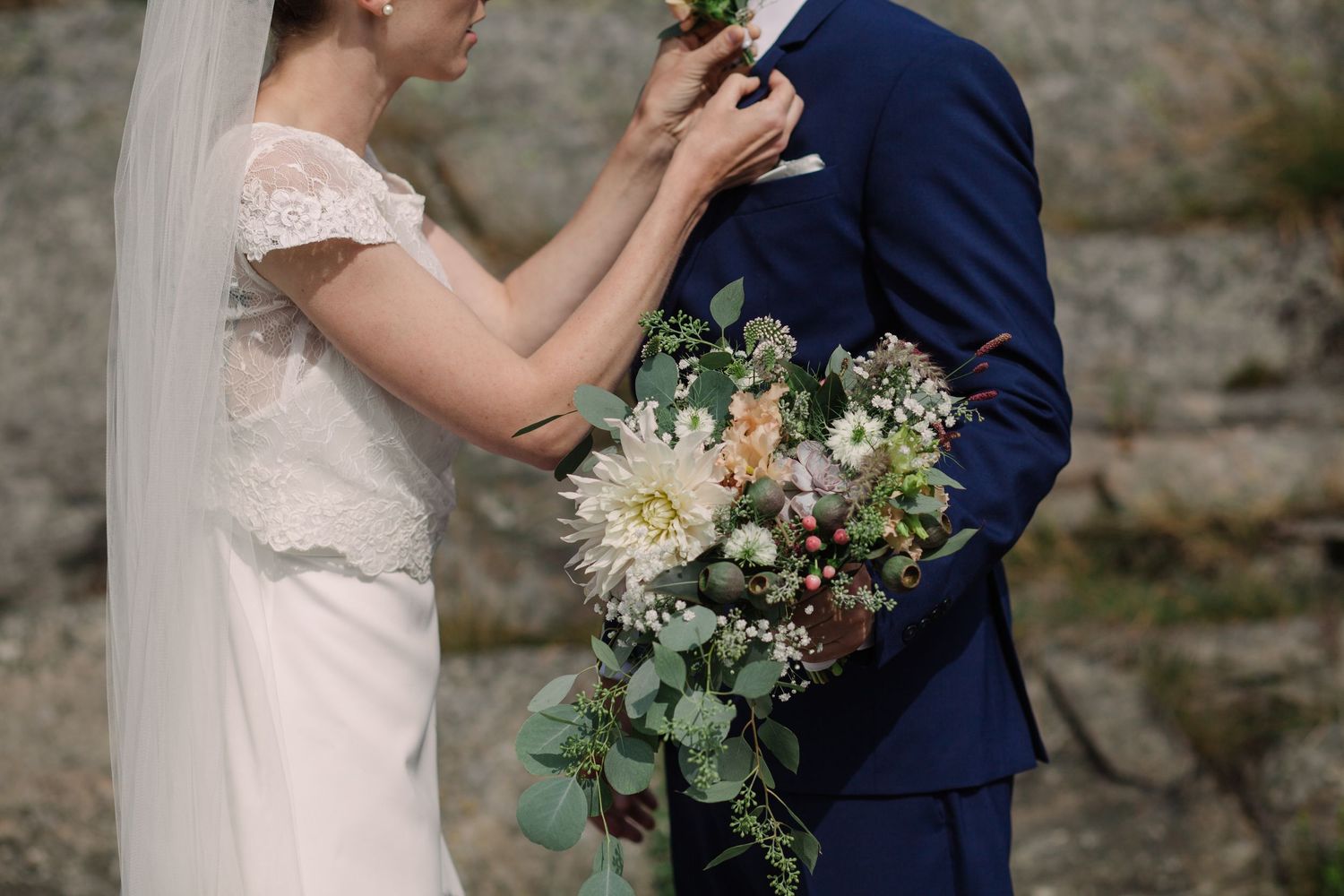 A bride in lace dress adjusts a boutonniere on a navy suit while holding a rustic white and green floral bouquet.