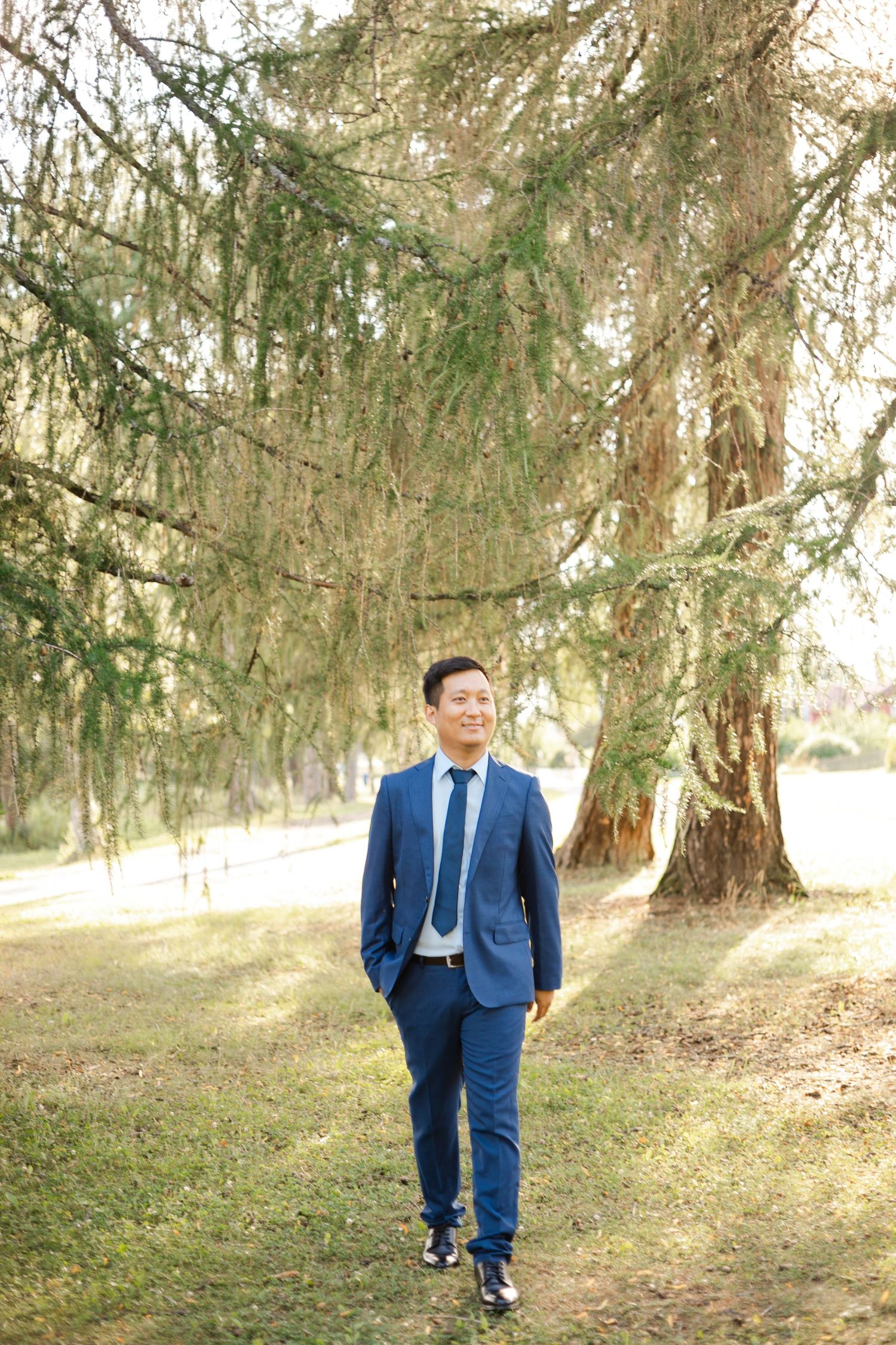 A person in a navy blue suit walks along a tree-lined path.