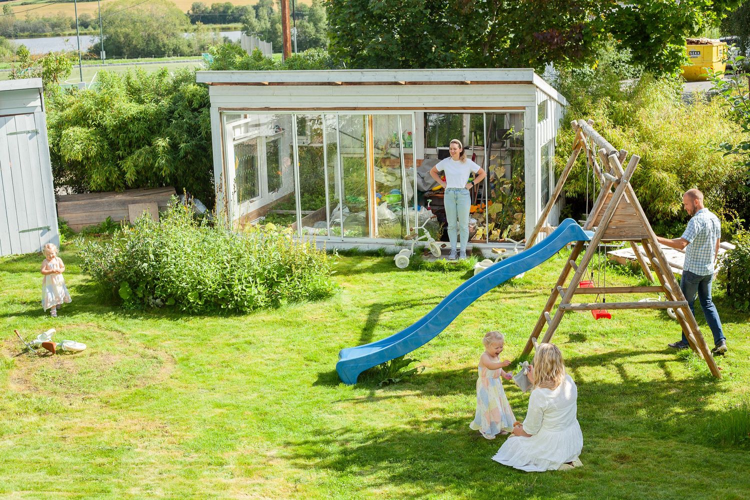 Children playing on a backyard playground with swings and slide near a greenhouse on a sunny day.