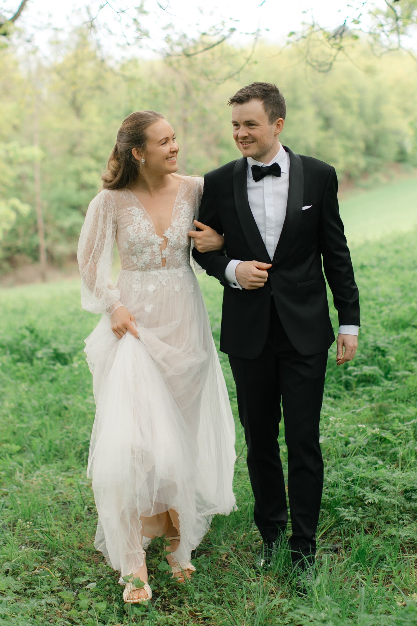 A bride in a white lace gown and groom in black tuxedo walk arm in arm through a lush green garden.