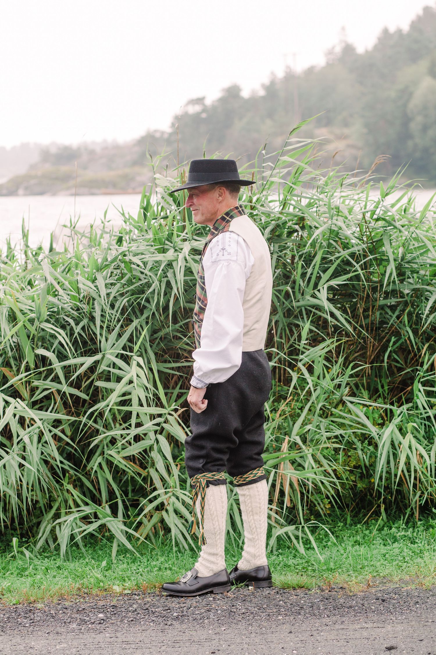 Person in formal attire and hat stands near tall grass on a misty coastal path.