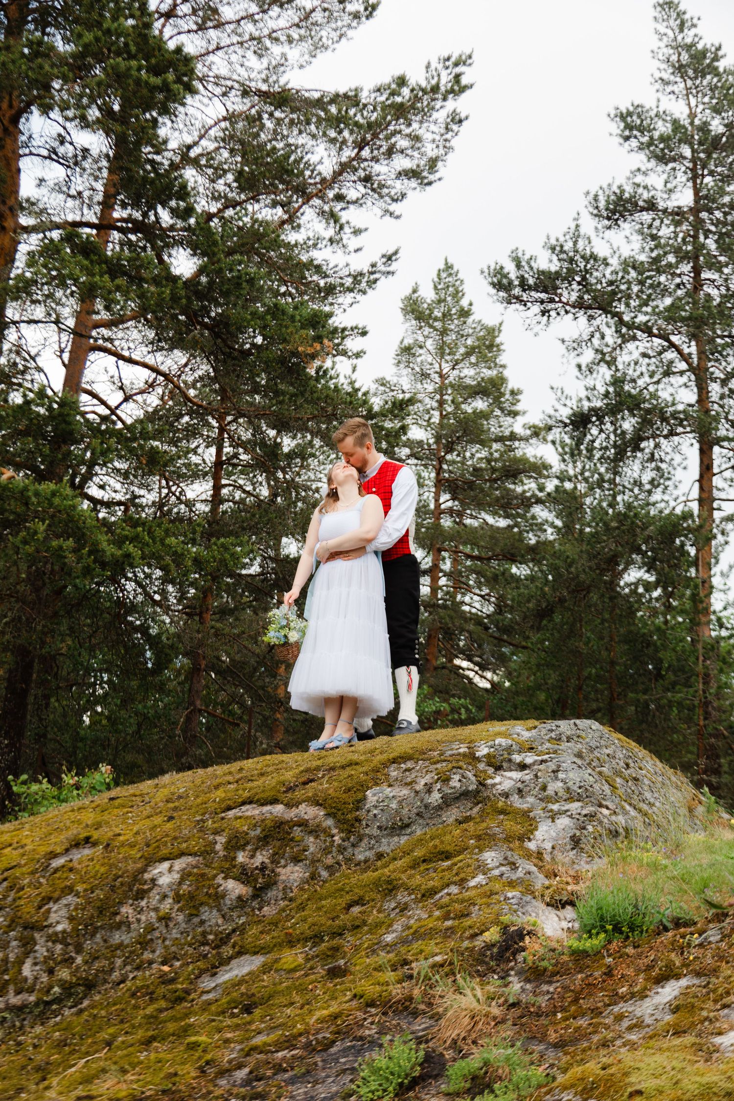 A couple in wedding attire stands on a scenic mossy rock overlooking a pine forest.