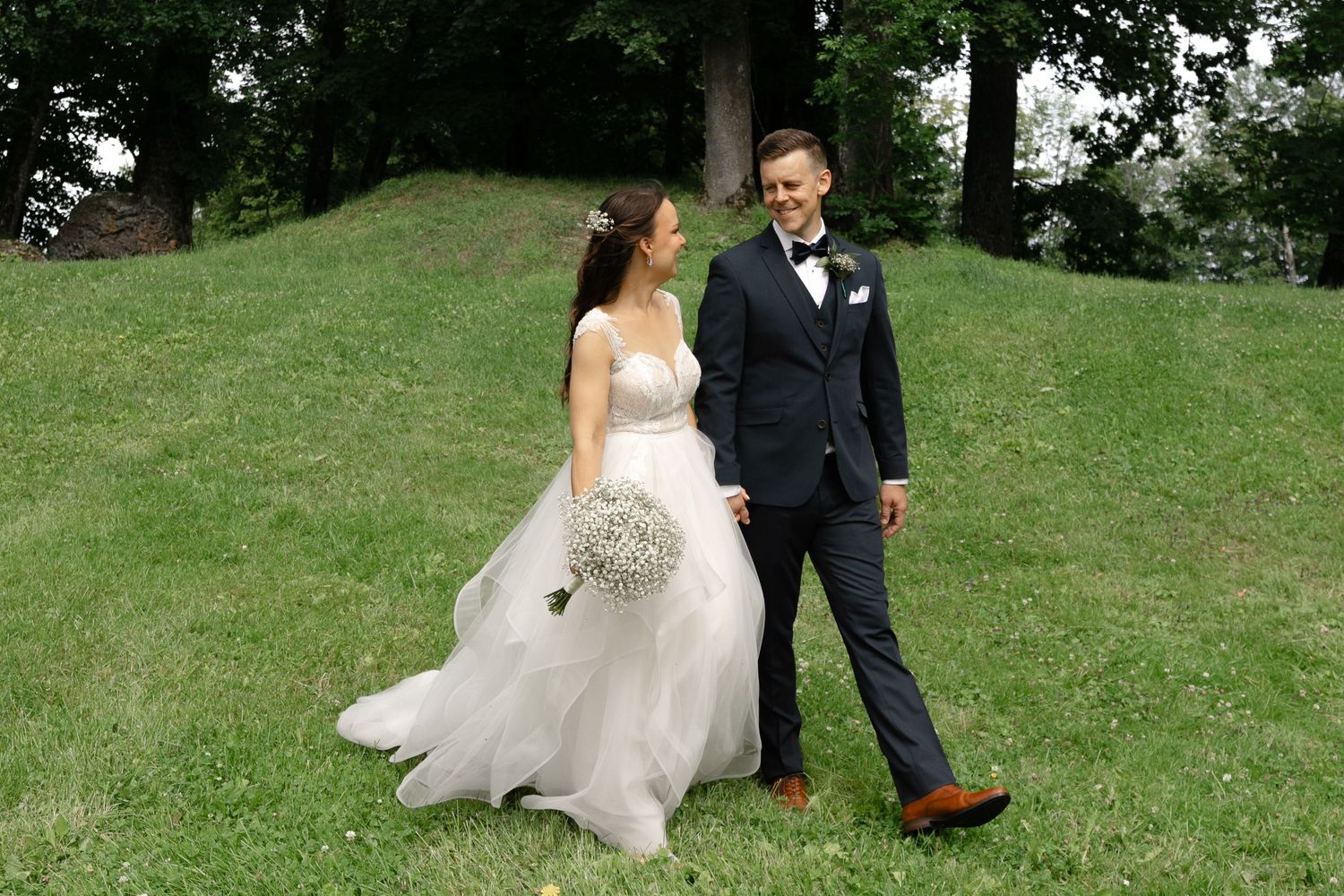 Wedding couple walking on a lush green lawn under tall trees.