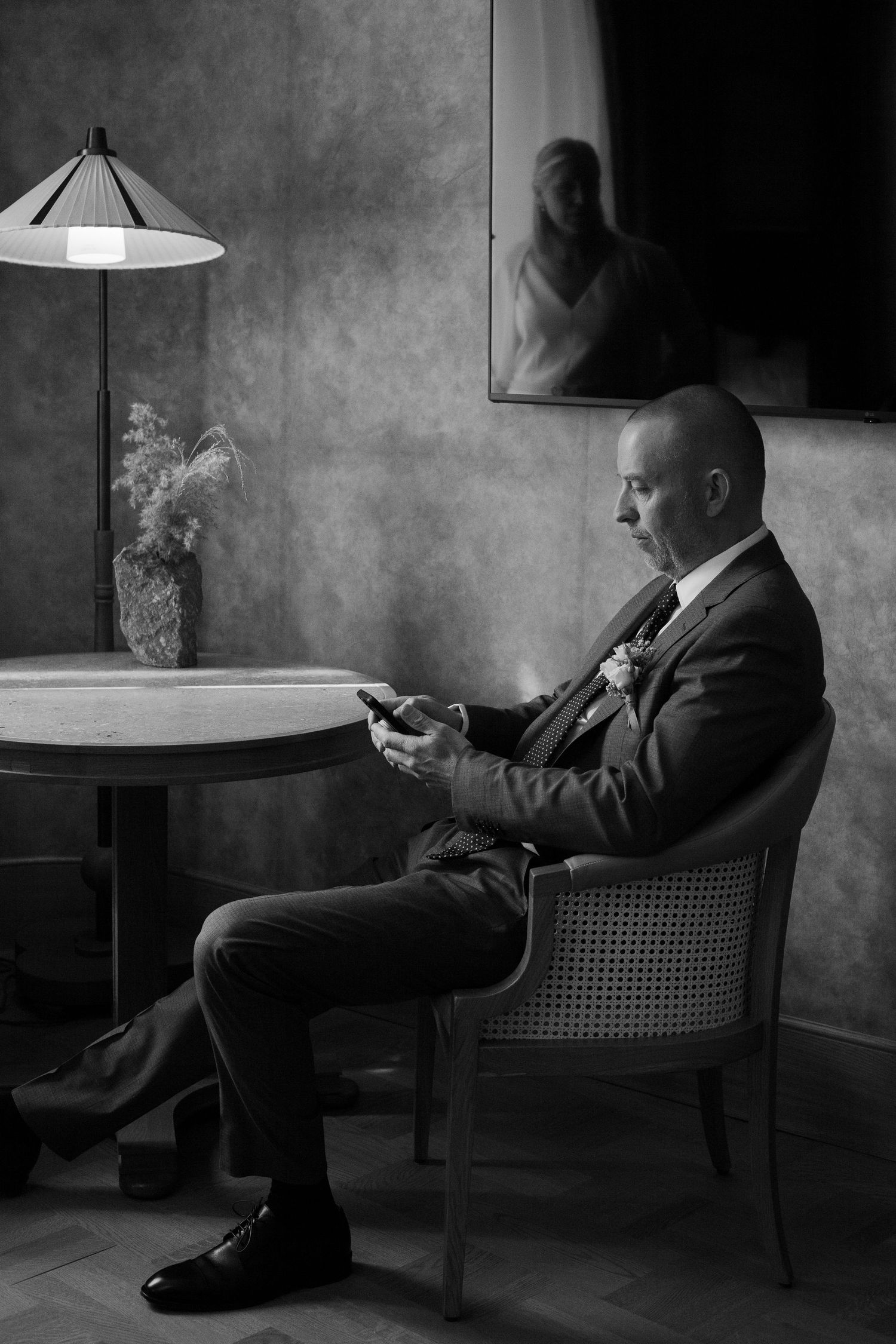 A person in formal attire relaxes in a cane-backed chair while reading in moody black and white lighting.