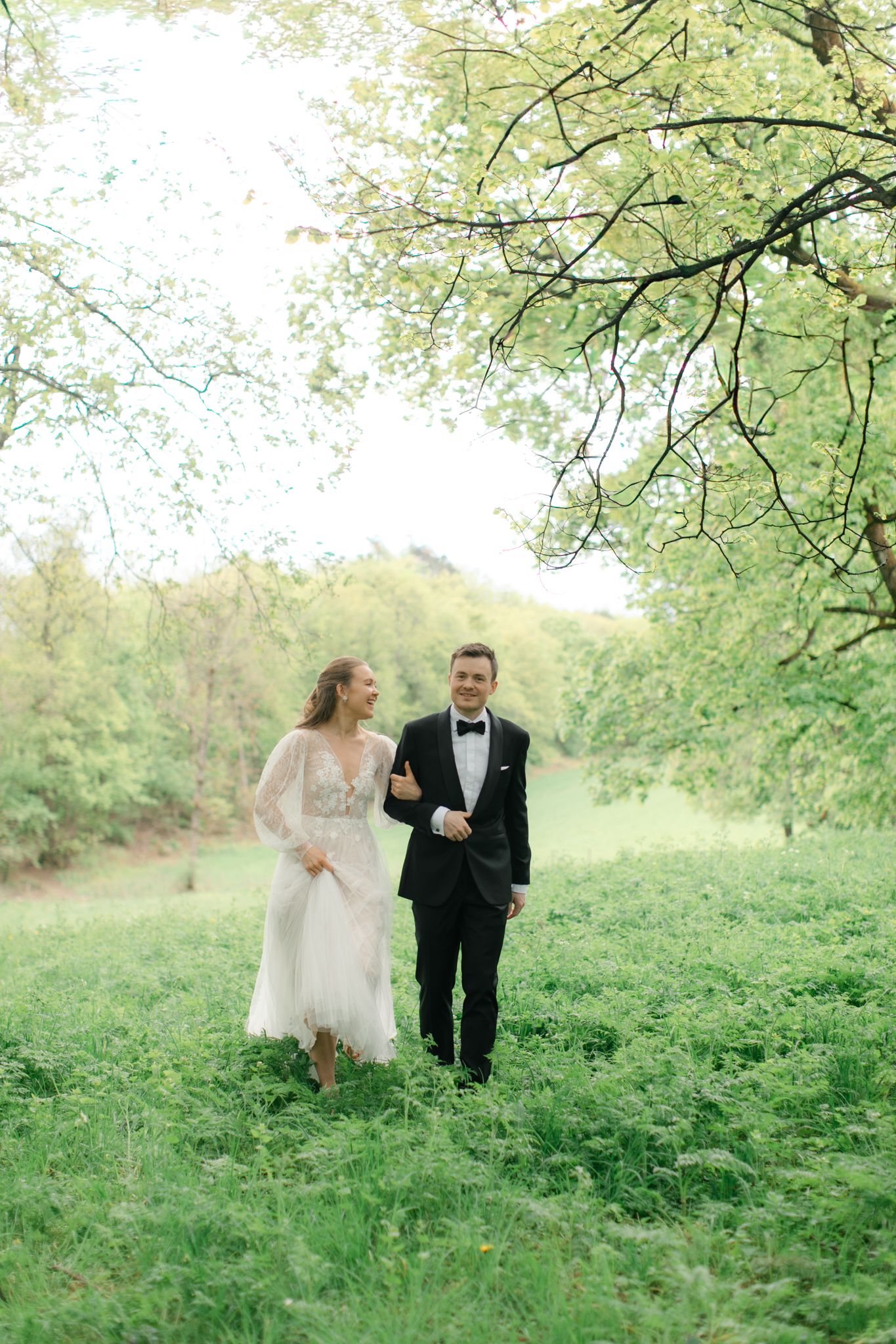 A bride and groom walk hand in hand through a lush green garden, smiling joyfully on their wedding day.