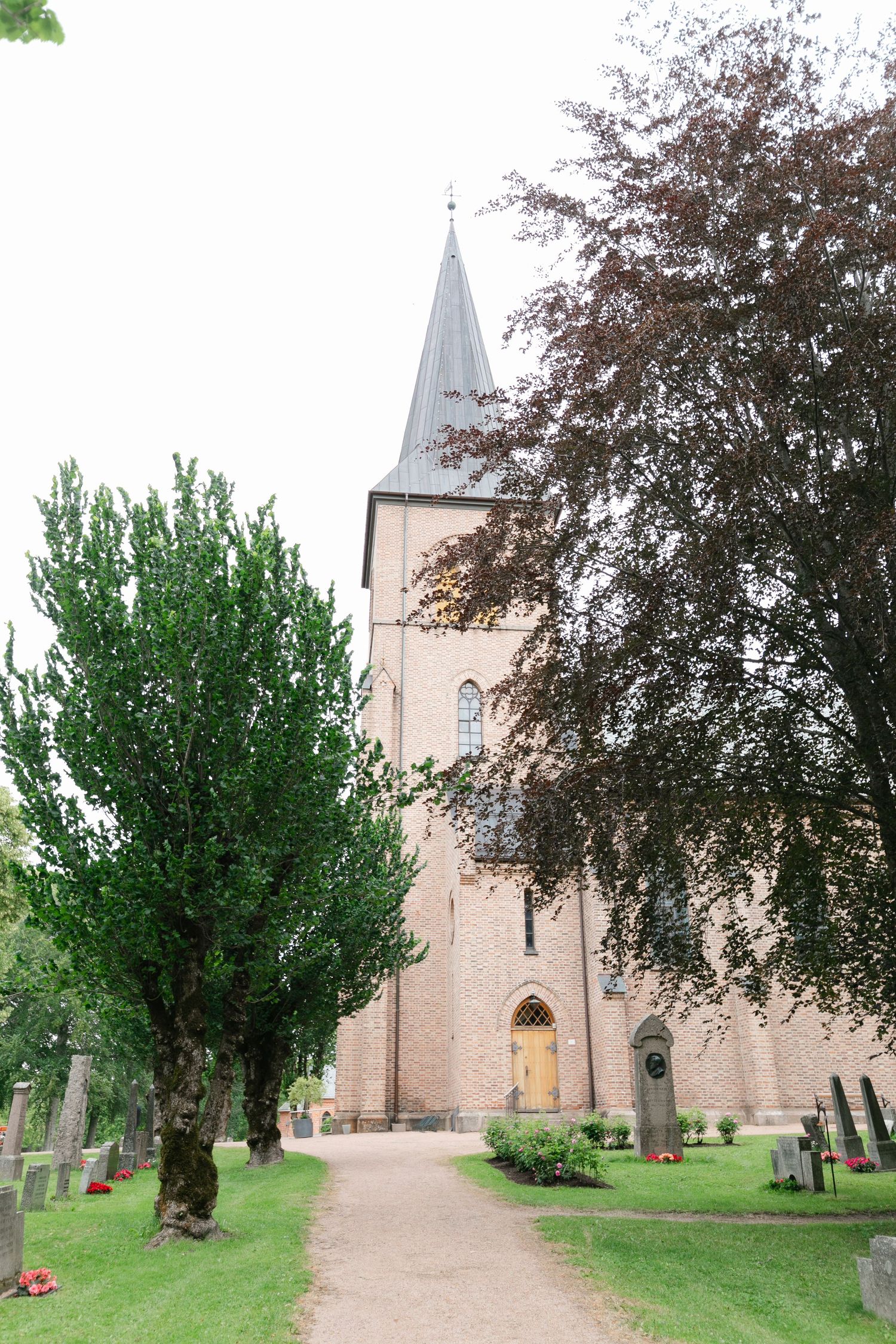 Tall church steeple and entrance framed by trees along a cemetery path.