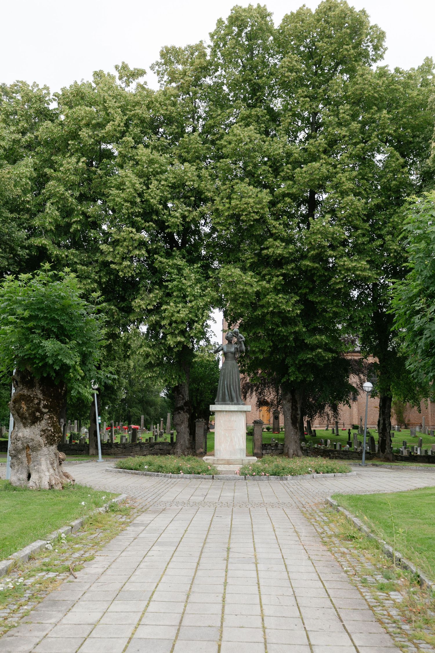 Stone statue centered on paved pathway surrounded by mature trees.