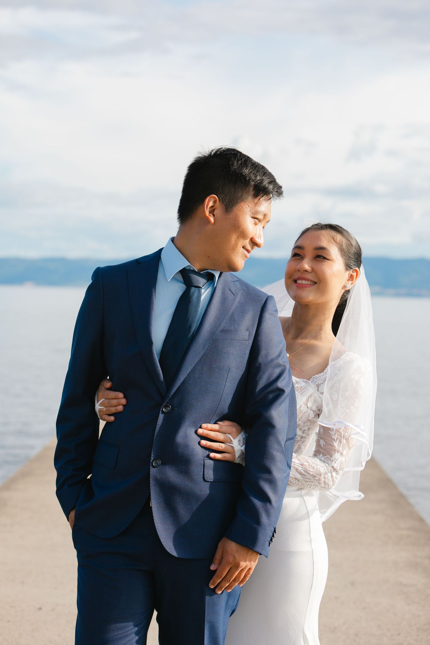 Newlyweds in formal attire sharing intimate moments on a pier by the water.