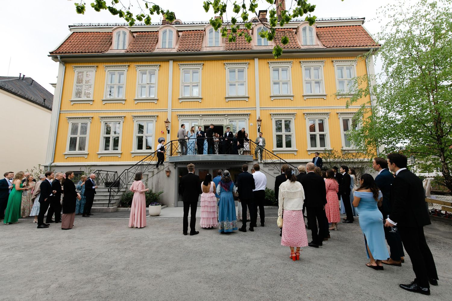 Elegant outdoor gathering at a yellow manor house with formally dressed guests on steps and courtyard.