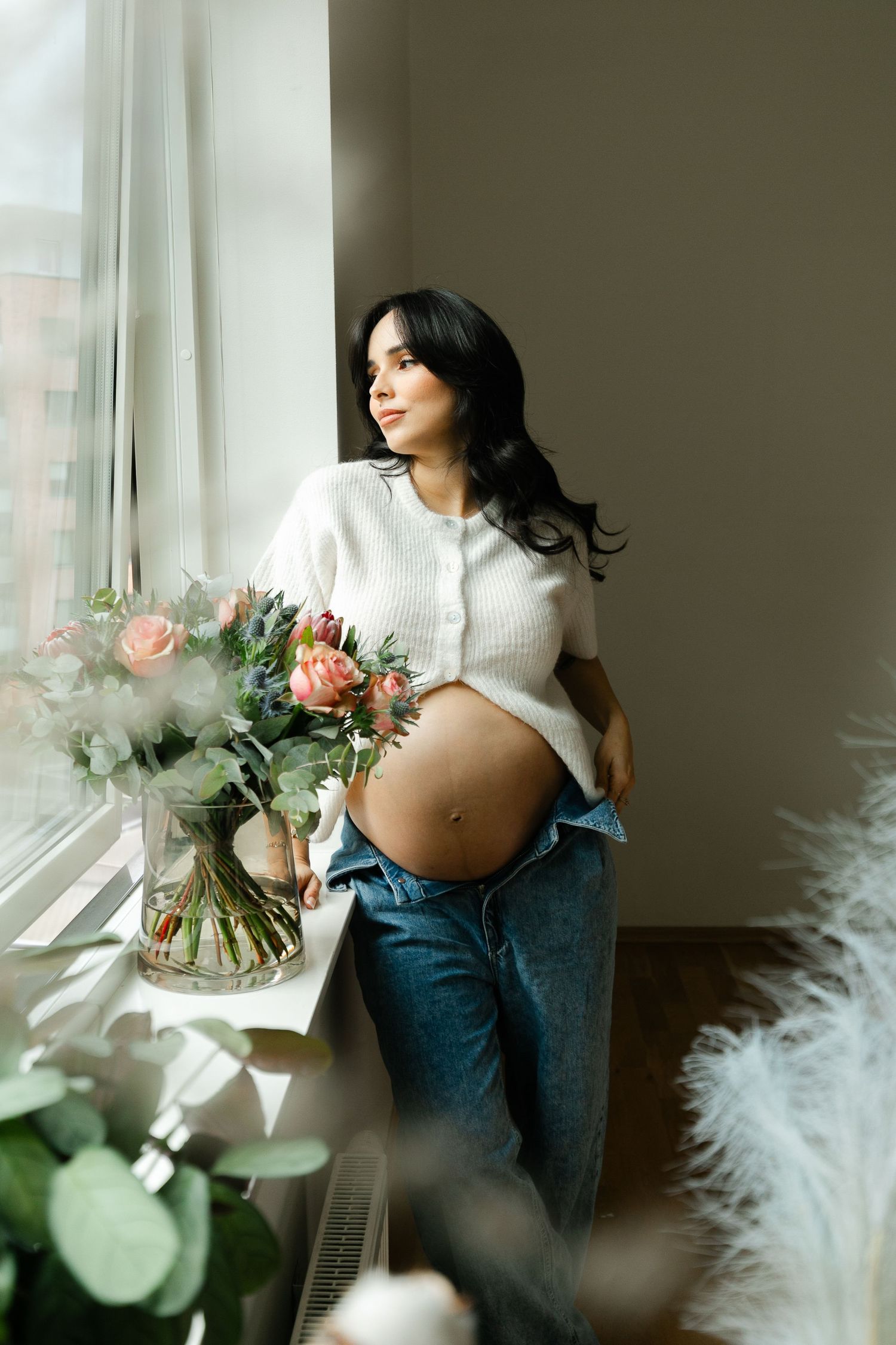 Maternity photoshoot featuring a white sweater, jeans, and floral arrangement by a sunlit window.