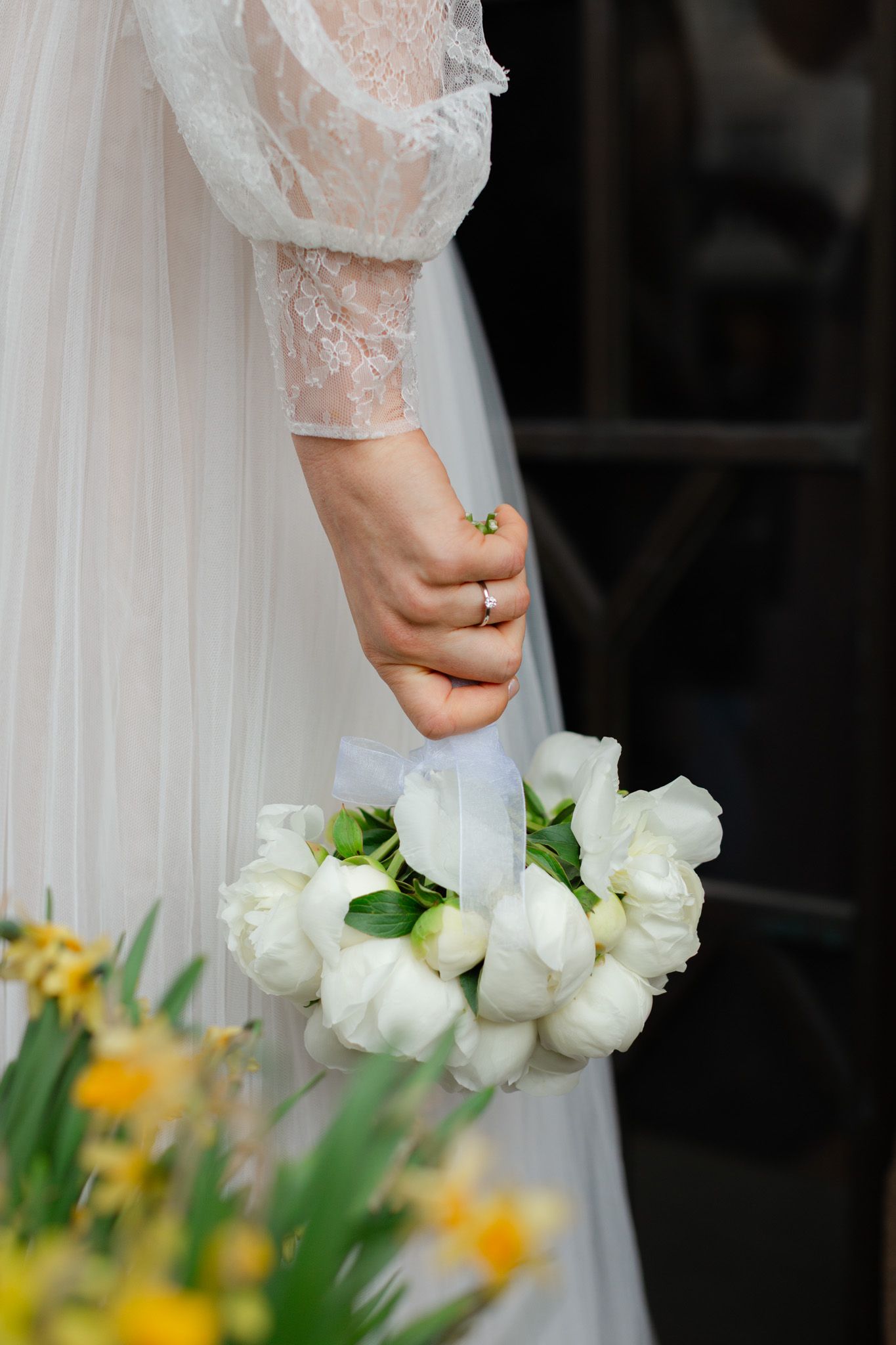 Bride in white lace gown holding elegant bouquet of white tulips with ribbon, ring visible on finger.