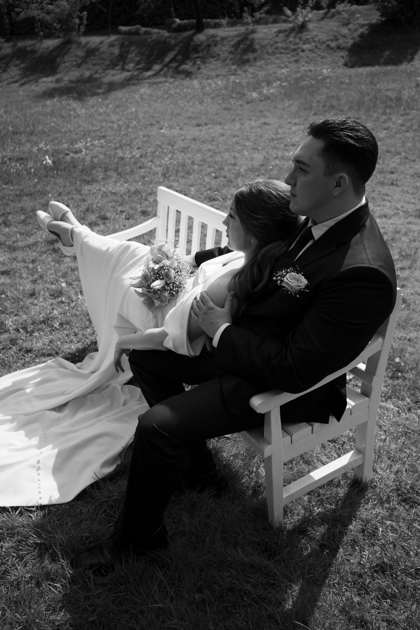Black and white photo of bride and groom sitting on a white bench, bride holding bouquet while resting in groom's arms.
