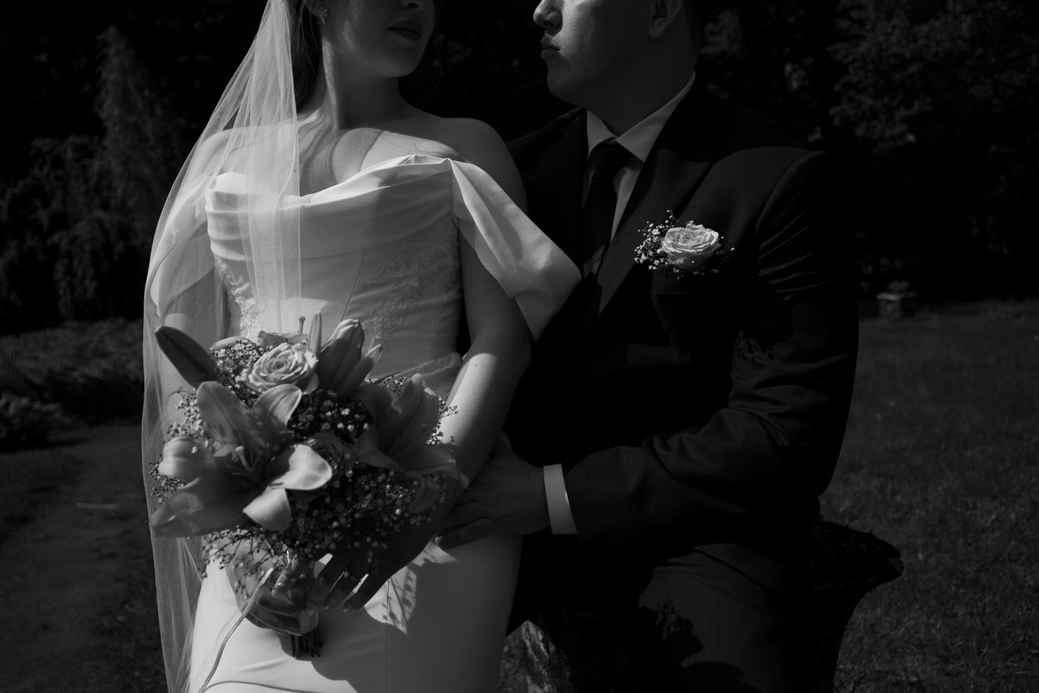 Black and white photo of a bride holding a bouquet and groom in a suit embracing outdoors at a wedding.