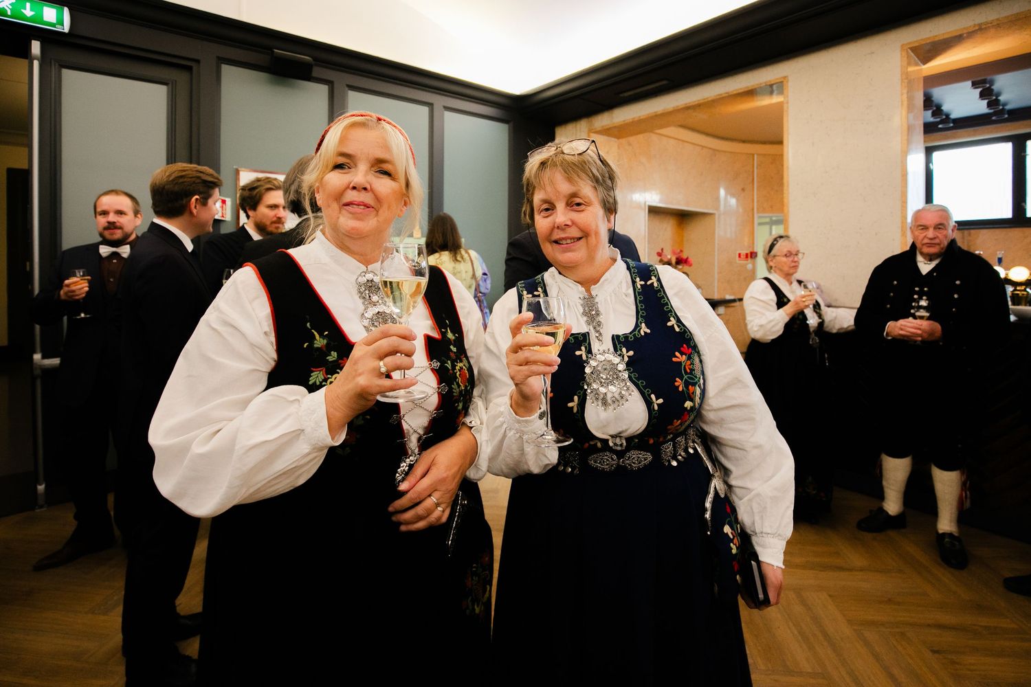 Two people in traditional dress toast with drinks at a celebration.