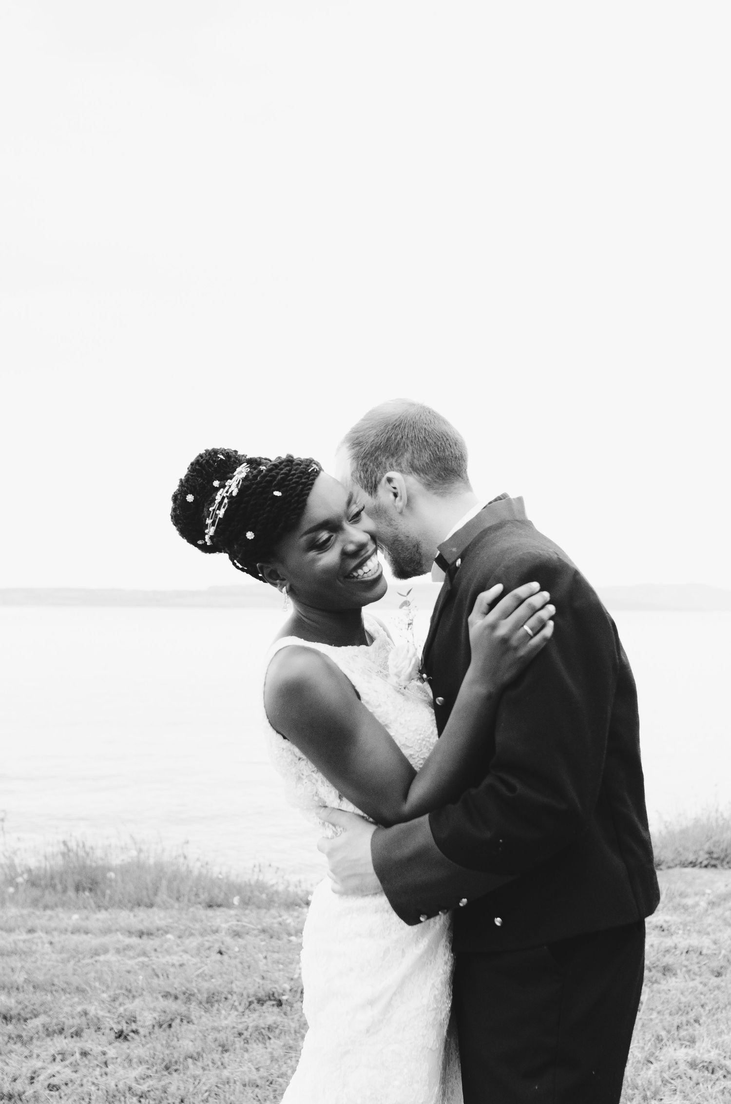 Black and white photo of a smiling bride and groom sharing a romantic moment outdoors near a waterfront.