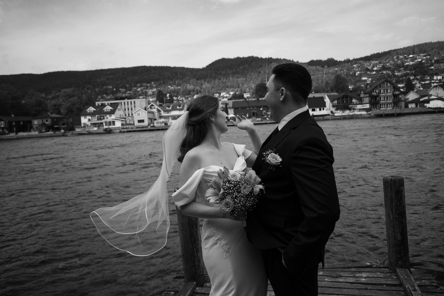 Black and white wedding photo of bride and groom on a lakeside dock with a European town and hills in the background.