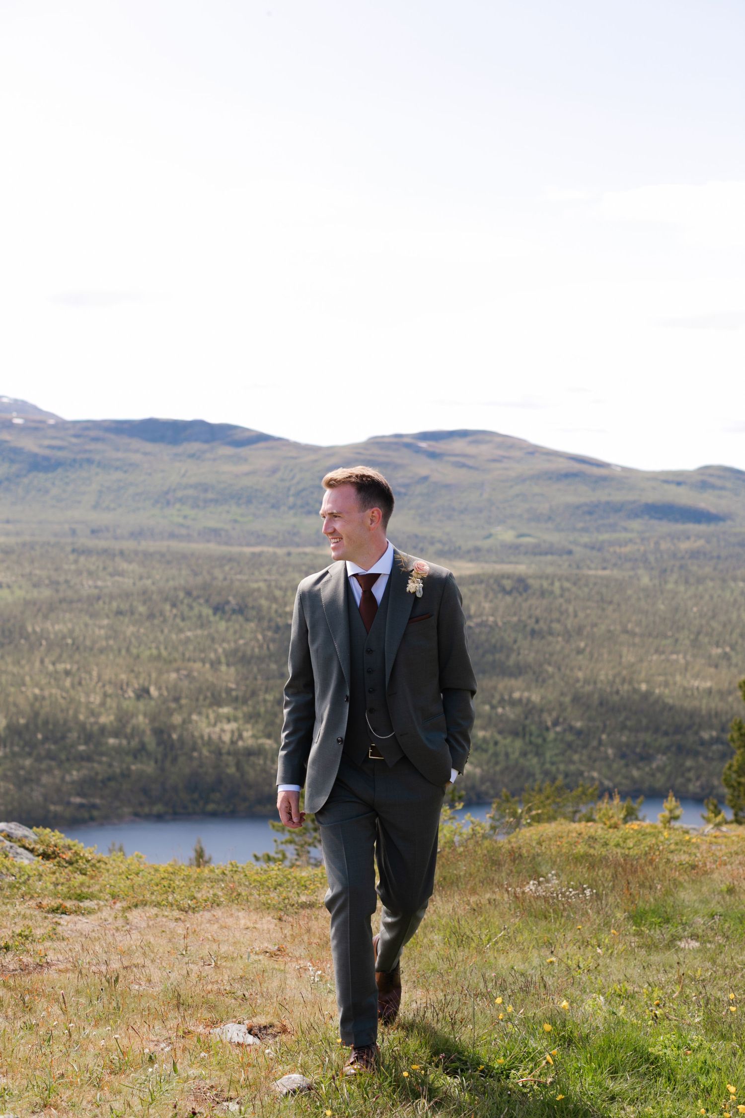 A couple walks along a scenic mountain hillside overlooking a lake and forest landscape.