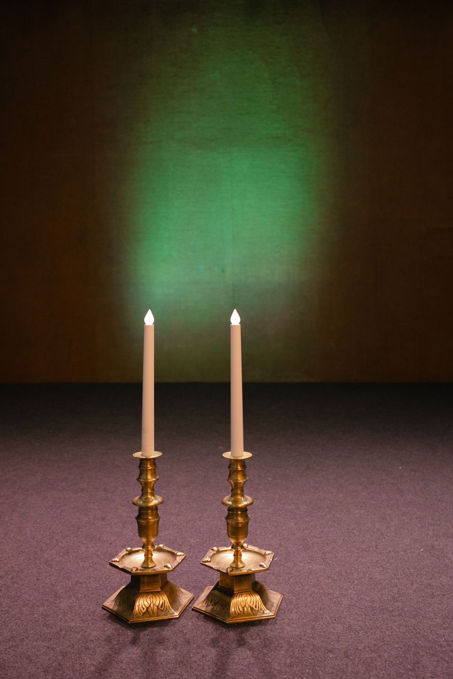 Two elegant brass candlesticks with white tapers against a dark background with green lighting.