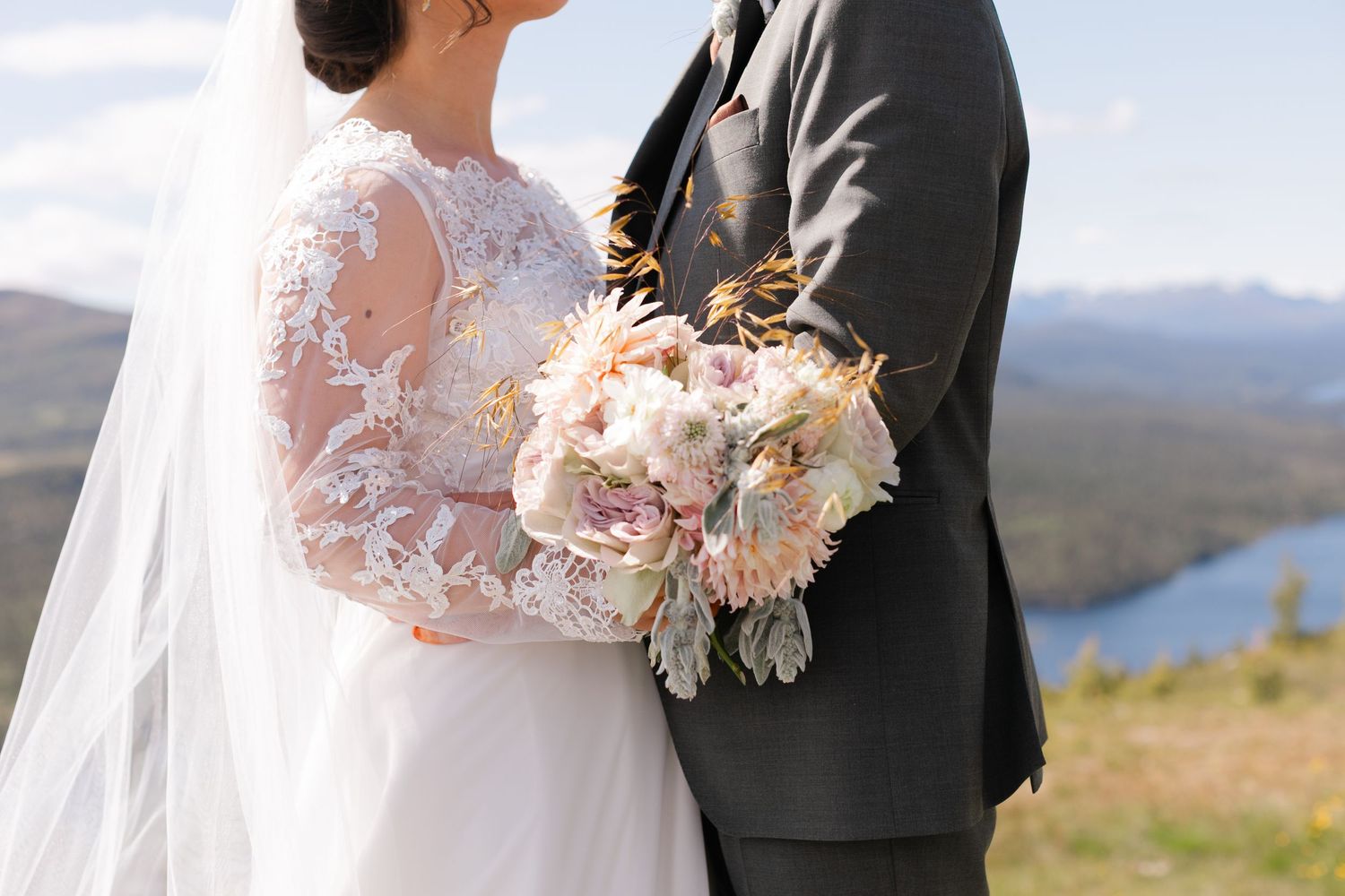 Bride and groom embrace on mountain top, bride holding blush peony bouquet with lace wedding dress and veil.