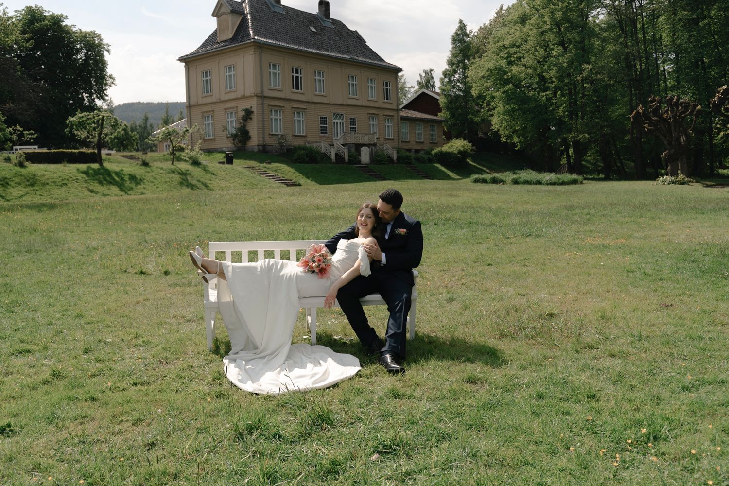 Bride and groom pose playfully on green lawn before elegant country estate on their wedding day.