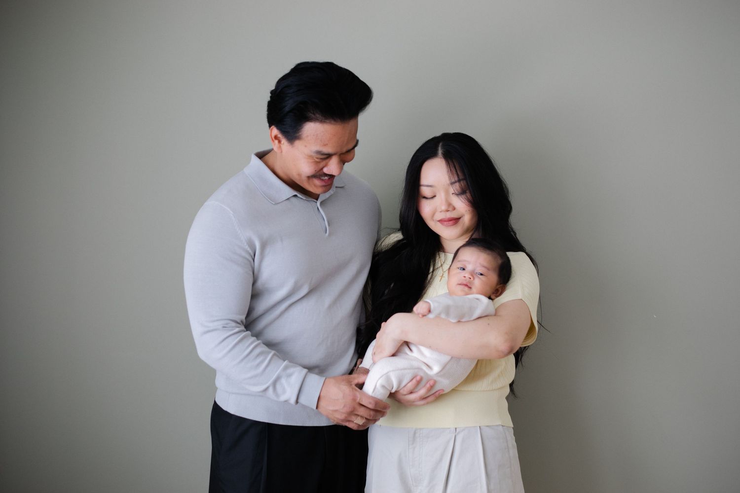 Family of three sharing a tender moment against grey wall background.
